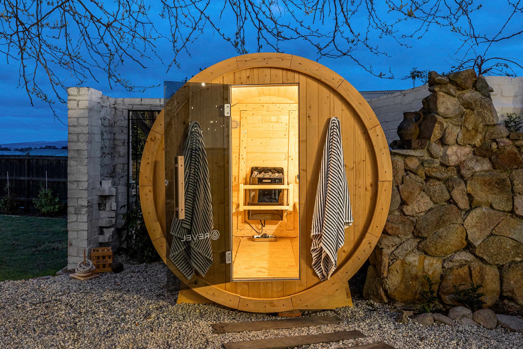 A barrel sauna glowing at dusk with towels hanging on the door