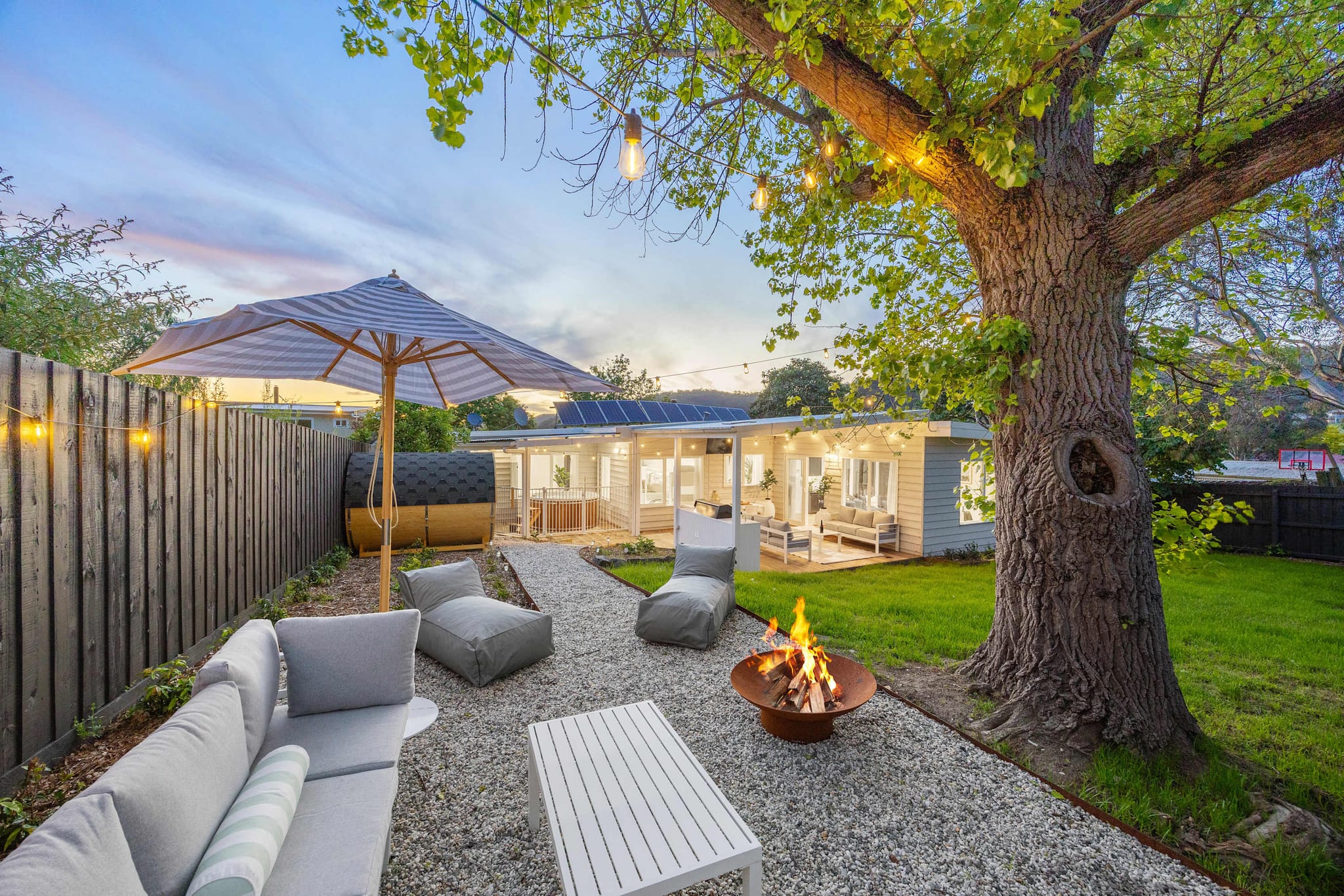A fire pit and lounge seating under a big tree at The Meadow at dusk, with fairy lights and the house glowing behind