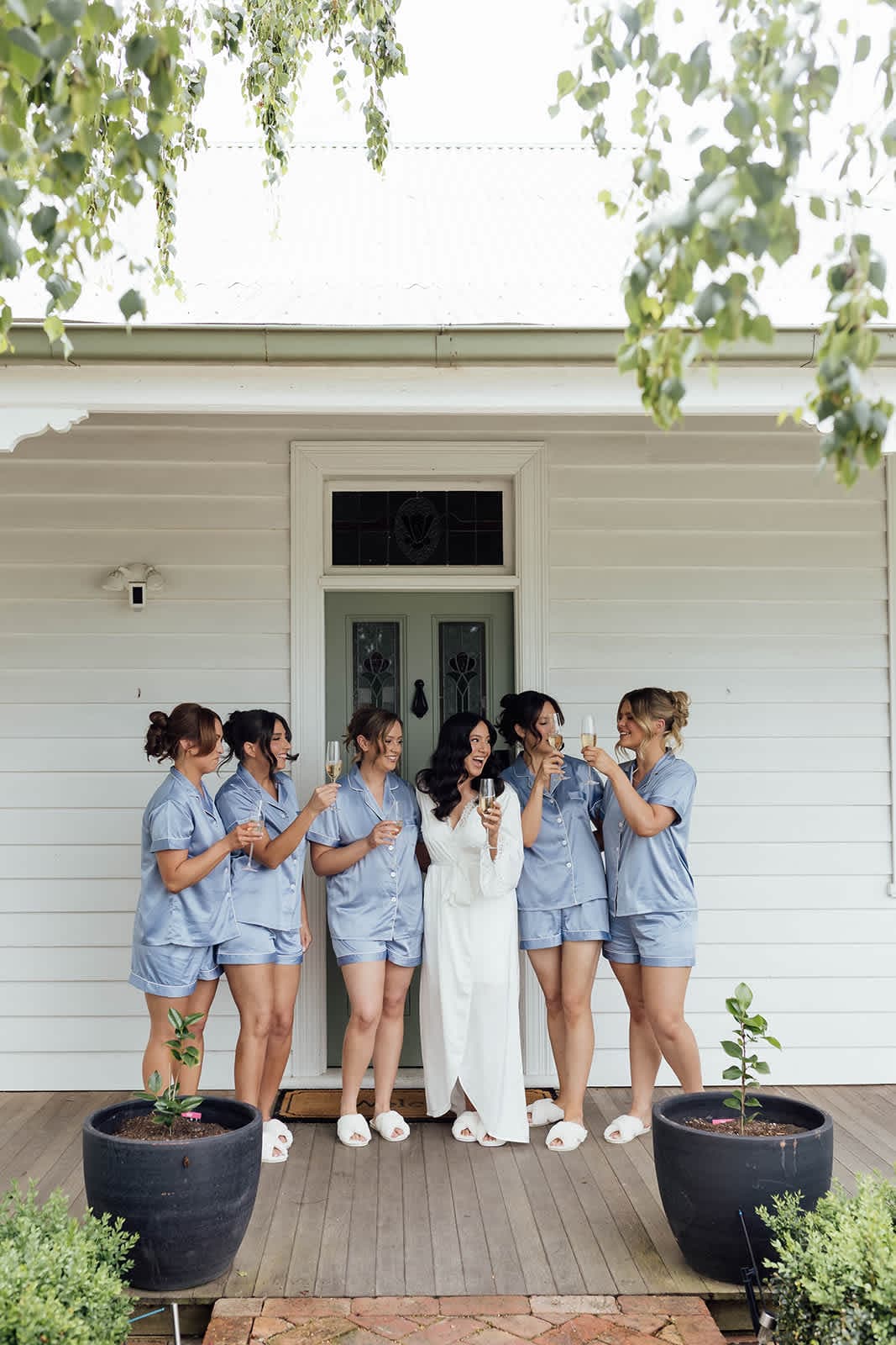 A bride in a white robe with her bridesmaids in matching pyjamas on the front porch, laughing