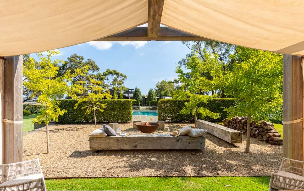 A fire bowl and day beds on gravel beneath a canvas pavilion at Musk Creek, with a hedge and the pool behind